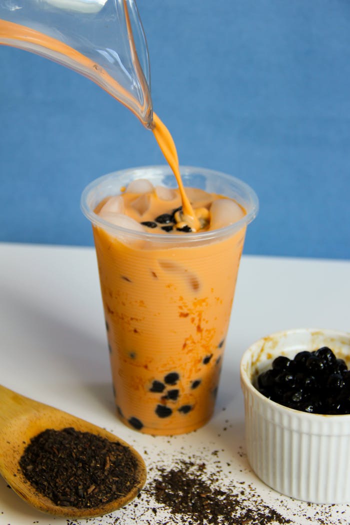 A fresh cup of Thai milk tea being poured, featuring black tapioca pearls and scattered tea leaves.